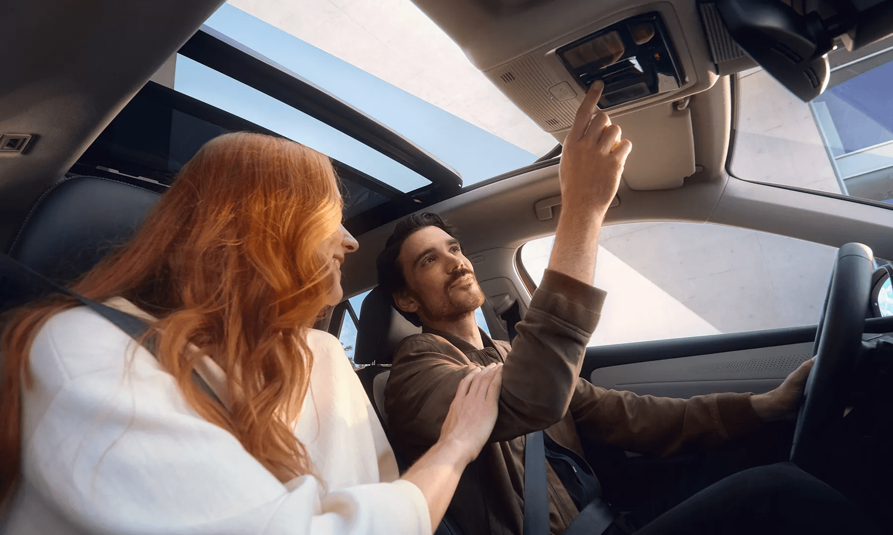 Pāris automobilī ar atvērtu panorāmas jumtu un saulainu debesu skatu.