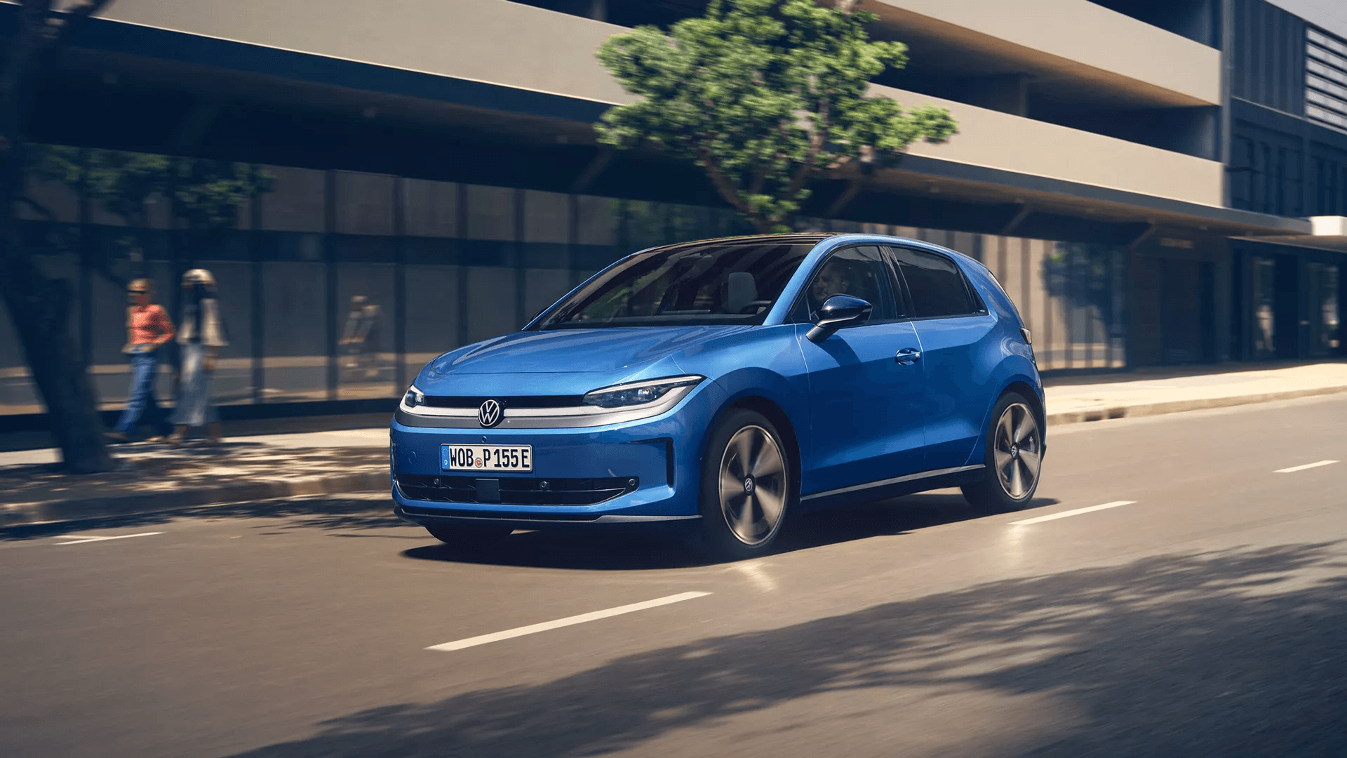 Gaiši zila Volkswagen automašīna brauc pa pilsētas ielu saulainā dienā. Fonā uz ietves redzami gājēji un ēku fasādes.