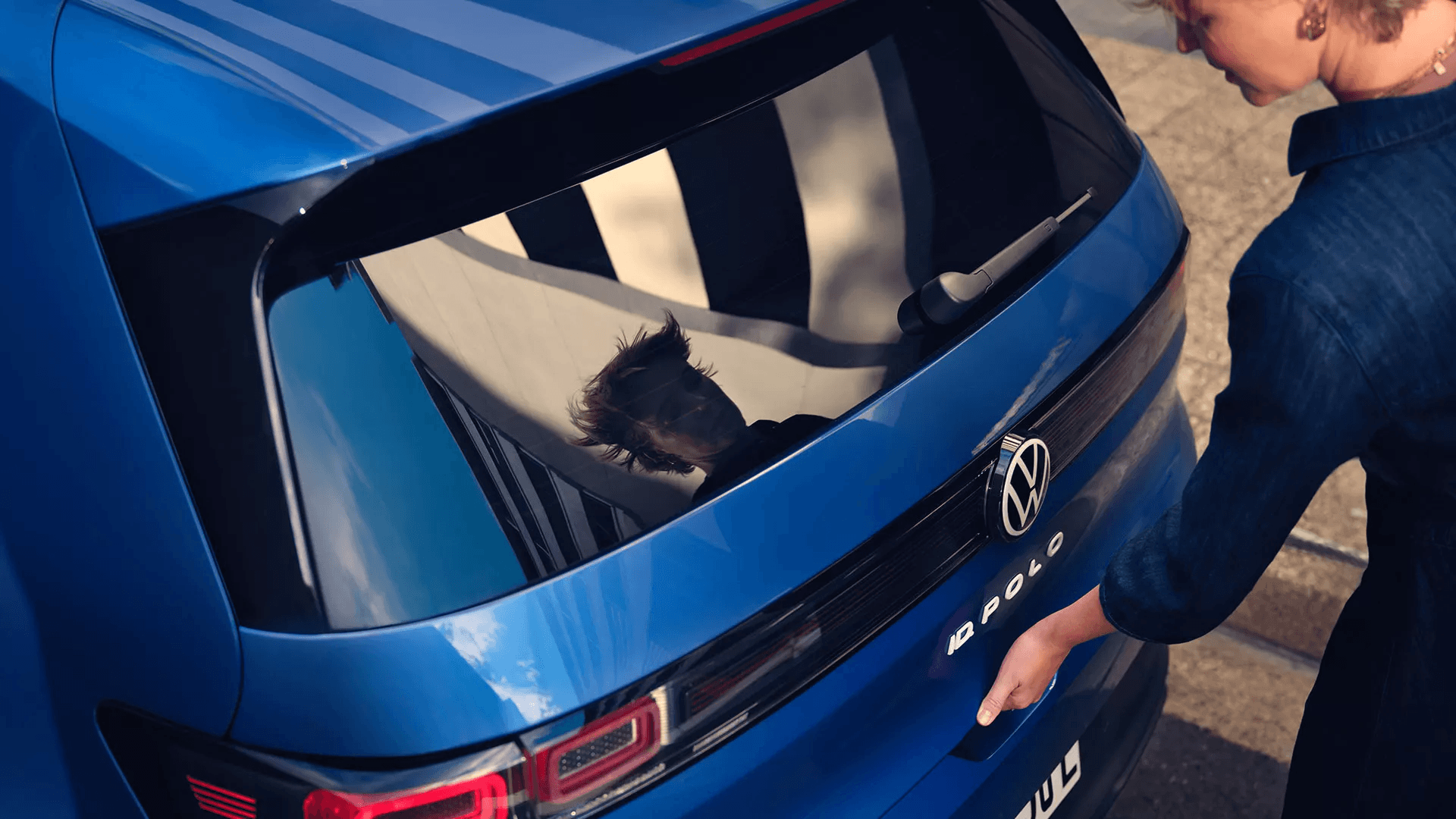Zilas Volkswagen ID. Polo automašīnas aizmugures tuvplāns. Sieviete, kurai redzama tikai roka un plecs, atver bagāžnieku, savukārt automašīnas tonētajā aizmugurējā stiklā ir skaidri redzams cilvēka atspulgs.