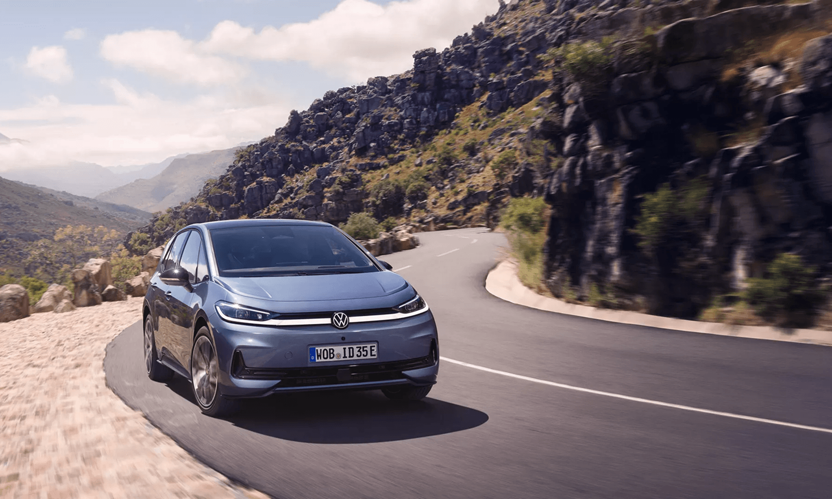 Zils Volkswagen ID.3 elektromobilis brauc pa līkumainu kalnu ceļu ar klinšainu ainavu fonā.