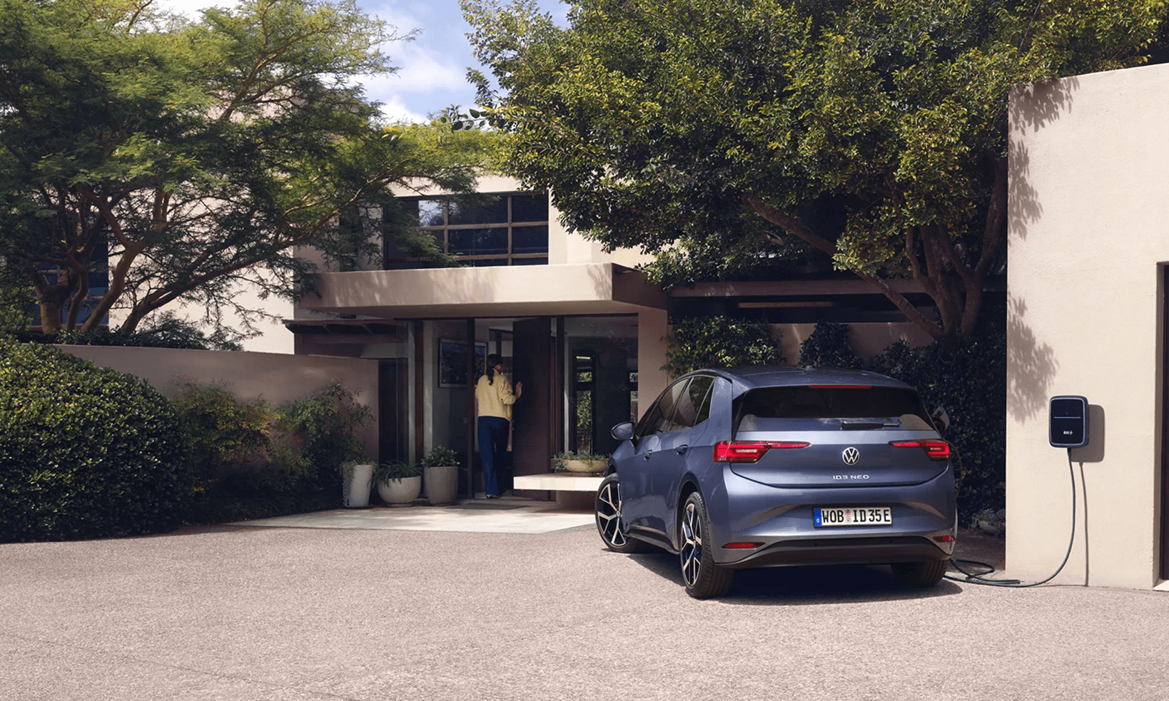 Zils Volkswagen ID.3 NEO elektromobilis uzlādējas mājas piebraucamajā ceļā, fonā sieviete ieiet modernā mājā.